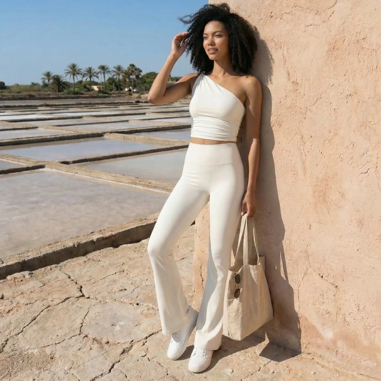 Une jeune femme à la chevelure bouclée pose devant un mur en terre cuite sous un soleil éclatant. Elle porte un ensemble immaculé composé d'un crop top asymétrique à une épaule et d'un legging évasé taille haute, complété par un sac en toile beige et des baskets blanches. L'arrière-plan révèle un paysage de salines et de palmiers sous un ciel bleu pur, évoquant une esthétique estivale et minimaliste.