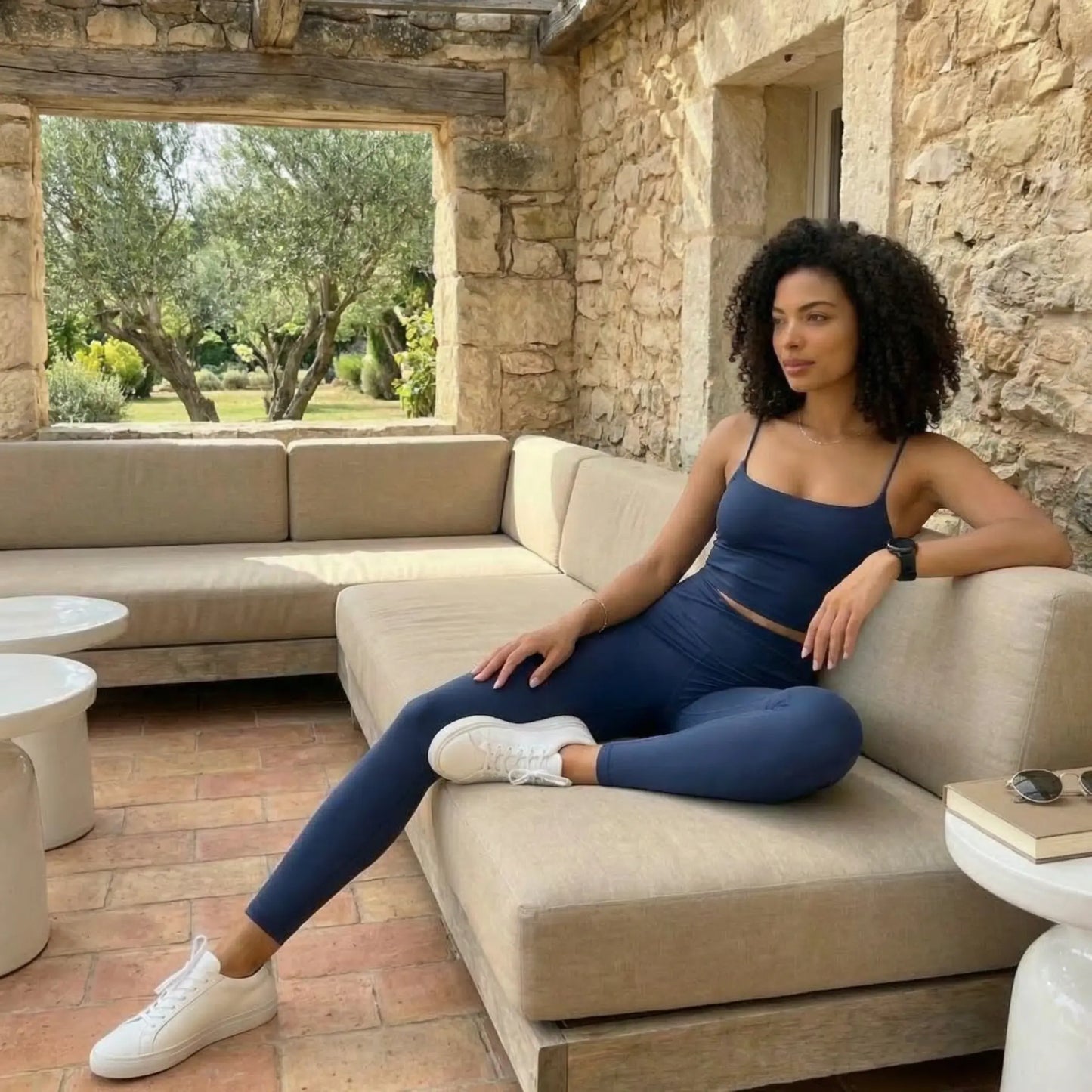 Une jeune femme aux cheveux bouclés pose élégamment sur un canapé d'extérieur beige dans un cadre rustique aux murs de pierres. Elle porte l'ensemble Elia bleu marine composé d'un legging taille haute et d'une brassière à fines bretelles, associé à des baskets blanches épurées.