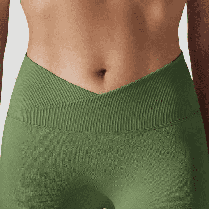 Cette image présente le legging Elia de Fyness Inc, mettant l'accent sur sa coupe innovante avec une taille haute croisée en V qui flatte la silhouette. Le coloris vert olive s'harmonise avec un arrière-plan estival composé de maisons méditerranéennes blanches et d'une eau bleu azur, soulignant l'aspect polyvalent et élégant du vêtement.