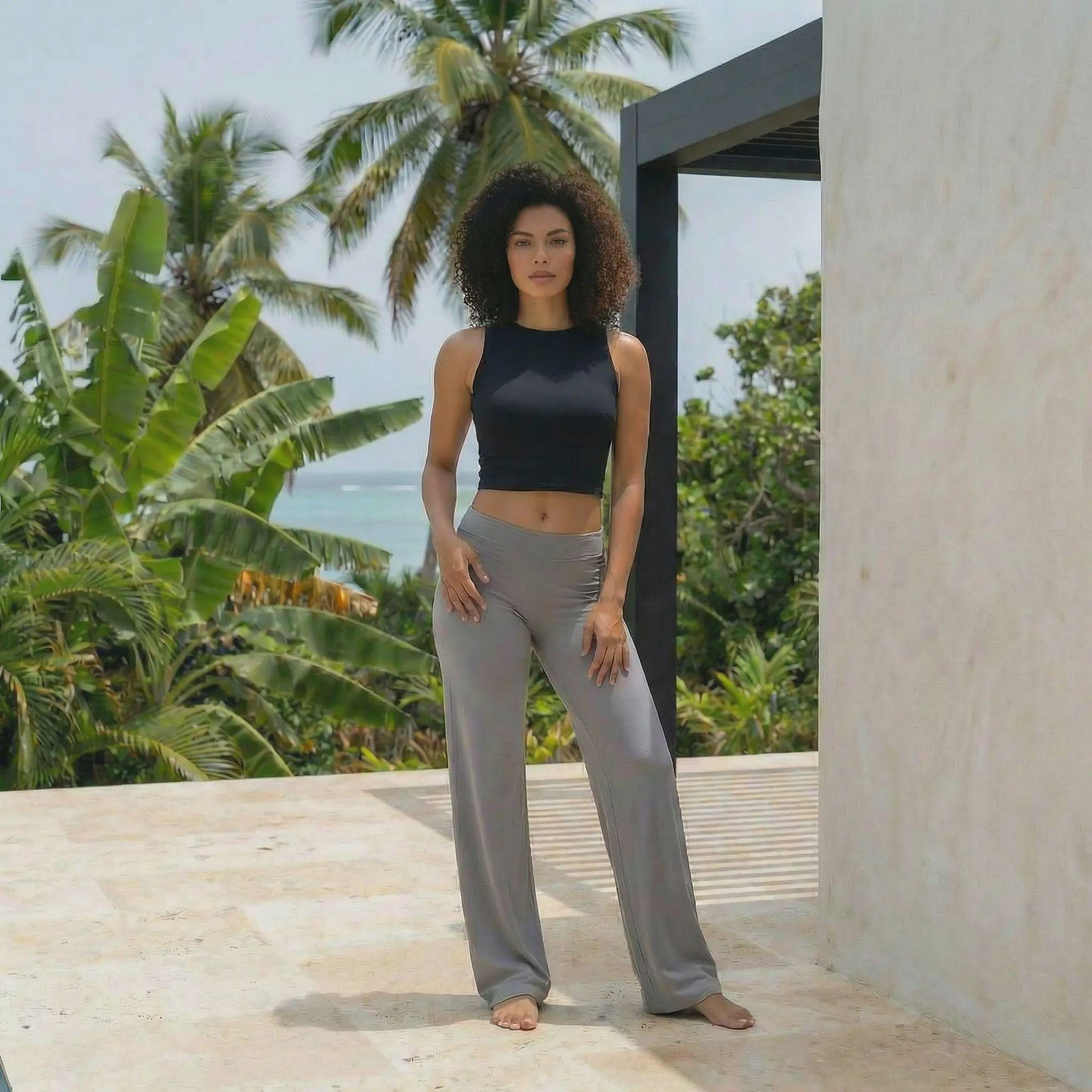 Une jeune femme aux cheveux bouclés pose sur une terrasse en pierre claire, vêtue d'un legging évasé gris taille haute et d'un crop top noir. L'arrière-plan luxuriant composé de palmiers et d'une vue sur l'océan souligne l'aspect confortable et polyvalent de cette tenue de sport et de détente.