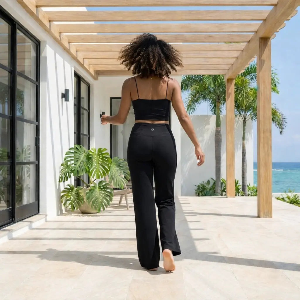 Une femme aux cheveux bouclés marche de dos sur une terrasse moderne et lumineuse, vêtue d'un ensemble de sport noir comprenant un legging évasé et un caraco. La scène se déroule sous une pergola en bois avec une vue dégagée sur des palmiers et l'océan turquoise en arrière-plan.