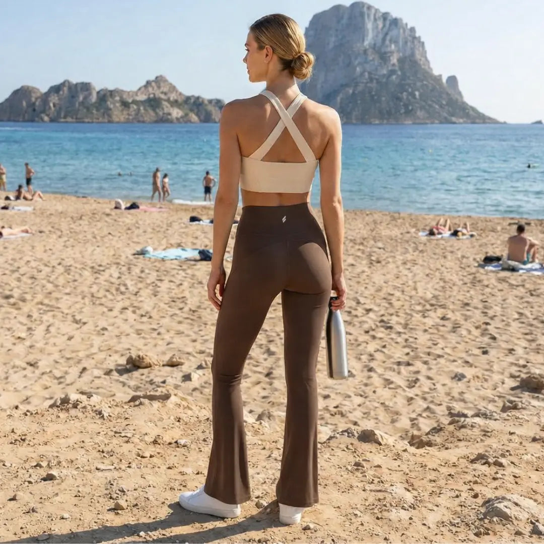 Une femme blonde vue de dos porte un legging de sport évasé marron taille haute associé à une brassière beige à bretelles croisées. Elle se tient debout sur une plage de sable, tenant une gourde en métal argenté face à un paysage côtier méditerranéen avec des falaises rocheuses et une eau turquoise.