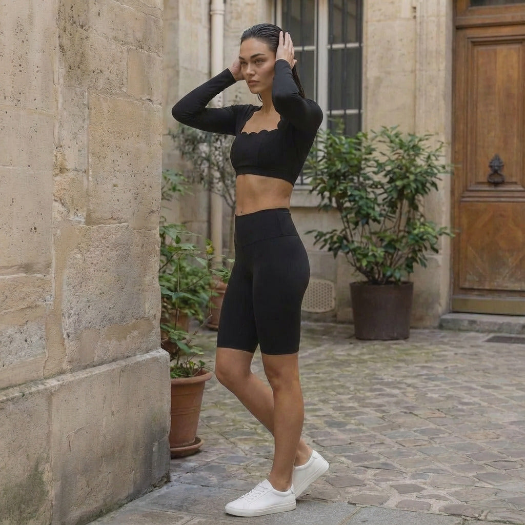 Une jeune femme pose de profil dans une cour intérieure au charme européen avec des murs en pierre de taille et un sol pavé. Elle porte un ensemble de sport noir élégant comprenant un top court à manches longues avec une encolure carrée aux bords festonnés et un short de cyclisme taille haute. Elle porte également des baskets blanches minimalistes. Ses cheveux bruns sont coiffés vers l'arrière avec un effet mouillé, ses mains posées délicatement sur sa nuque.