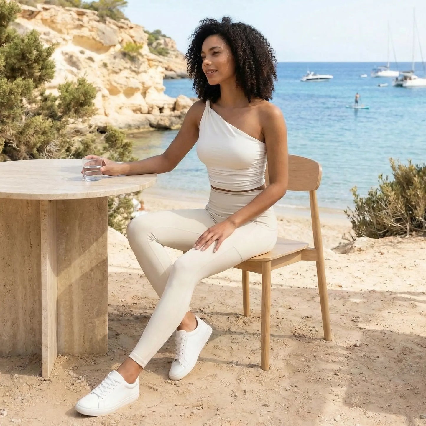 Une femme pose dans un cadre côtier idyllique, vêtue d'un ensemble de sport blanc de la marque Fyness composé d'un legging gainant et d'un haut asymétrique. Elle est assise sur une chaise en bois à une table en pierre, avec en arrière-plan une mer turquoise, des falaises et des voiliers sous un ciel clair.