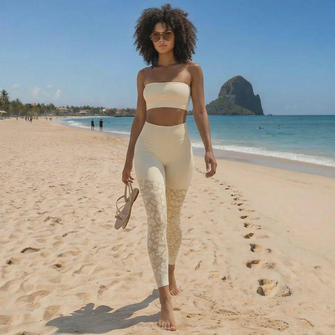 Une femme à la chevelure bouclée et lunettes de soleil marche sur une plage de sable fin, tenant ses sandales à la main. Elle porte un ensemble sport chic de couleur crème composé d'un bandeau et d'un legging galbant taille haute orné de motifs serpentins ou dentelle sur les jambes, avec un rocher volcanique et l'océan en arrière-plan.